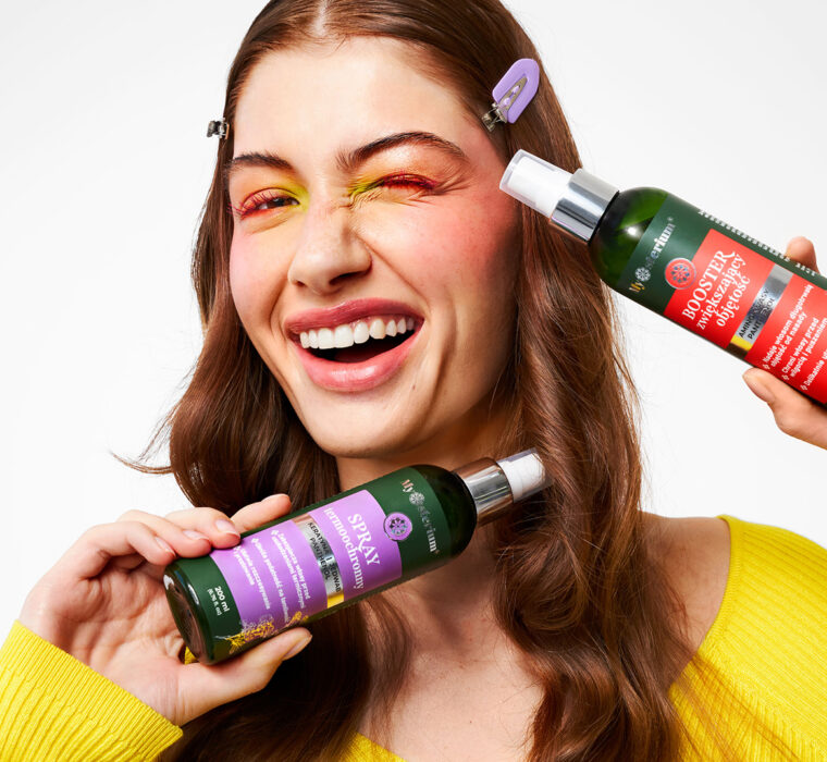 Uśmiechnięta kobieta prezentuje dwa kosmetyki do pielęgnacji włosów w sprayu.